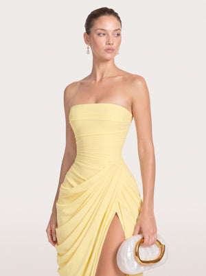 A woman modeling a strapless pale yellow ruched dress with a draped skirt and thigh-high slit, holding a white clutch with a gold ring handle against a light beige studio background.