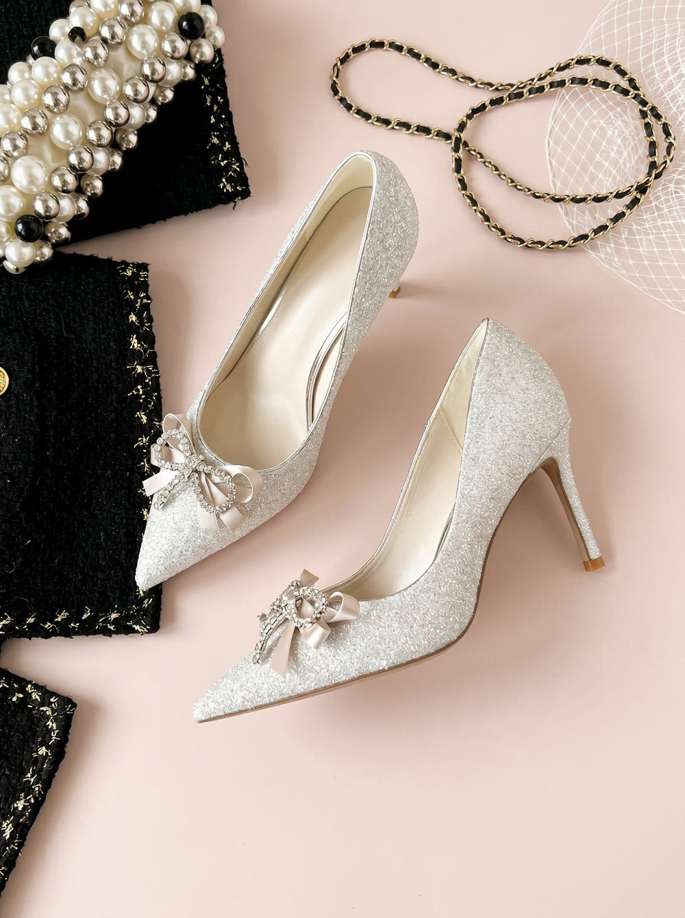 A single silver glitter pointed-toe pump with a slim high heel and a small pale pink bow embellishment on the toe, shown against a plain light beige studio background.